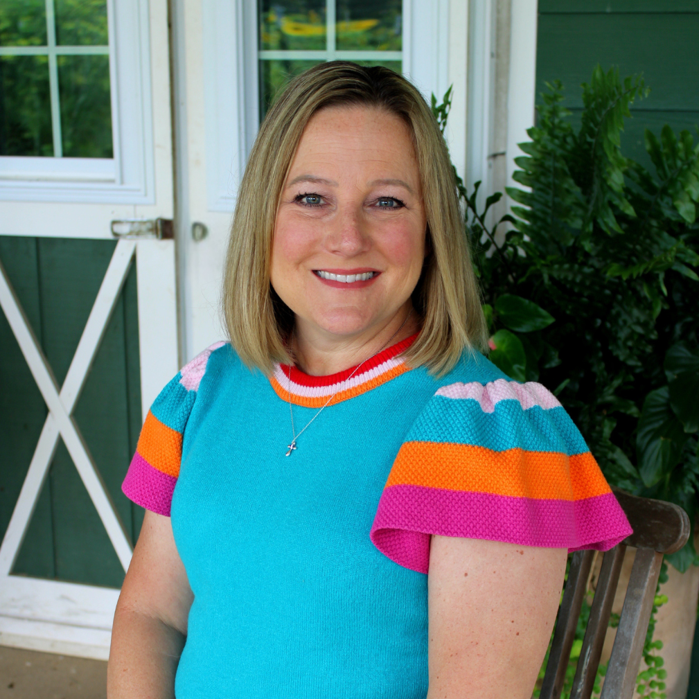 A smiling woman with blonde hair wearing a bright turquoise short-sleeve knit top with colorful striped accents on the collar and sleeves, sitting on a wooden chair on a porch.